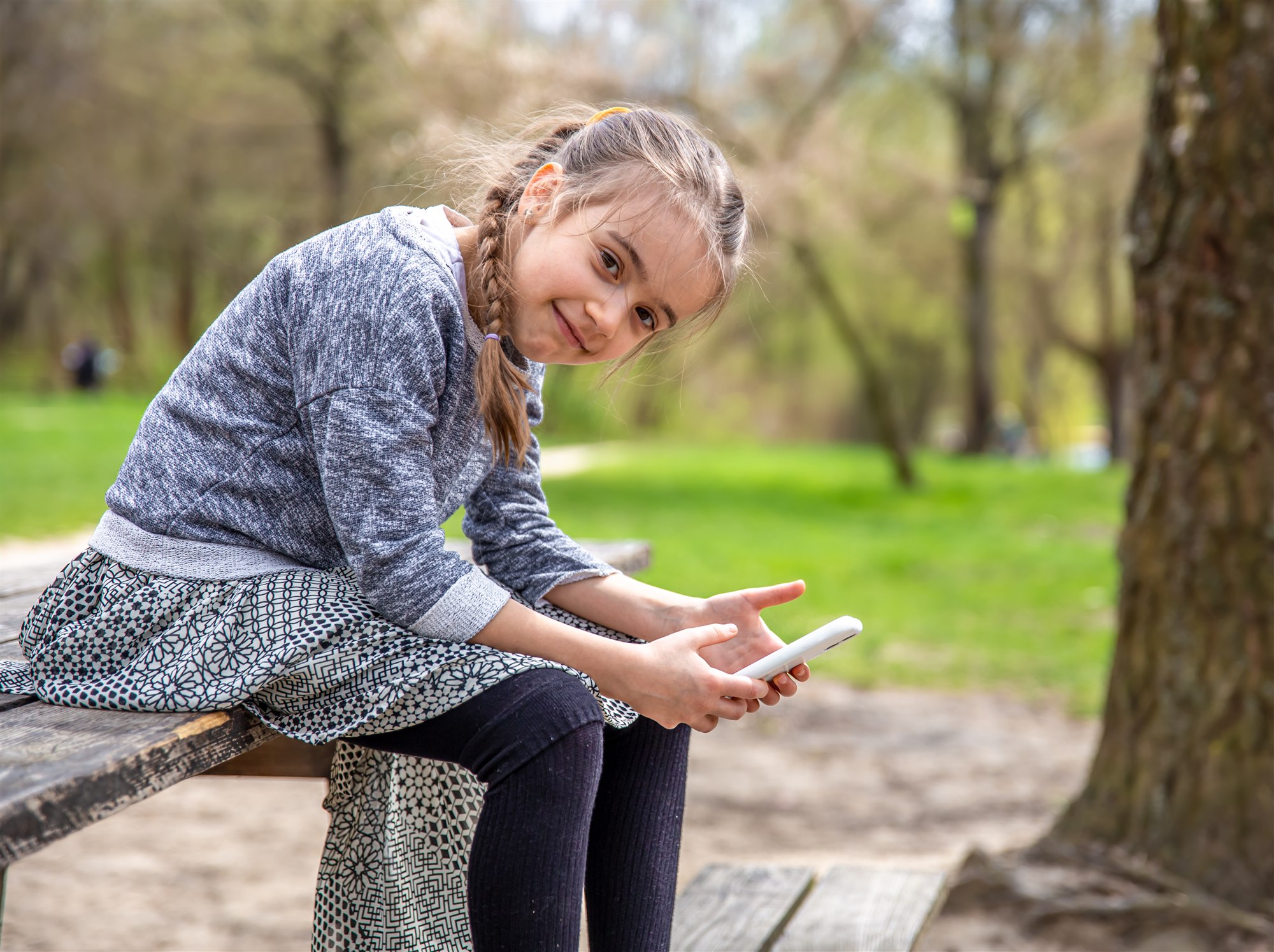 A child sitting on a park bench smiling while holding a phone